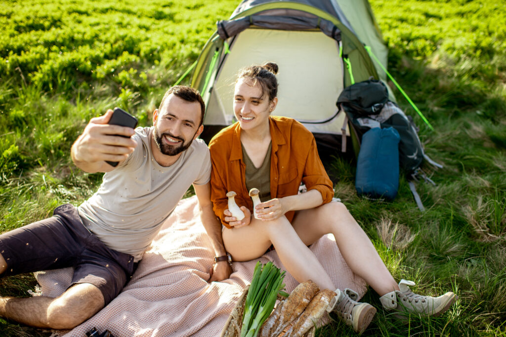 Smiling couple taking a selfie during a camping trip, sitting on a picnic blanket near their tent with fresh bread, vegetables, and camping gear, surrounded by lush green grass in a serene outdoor setting