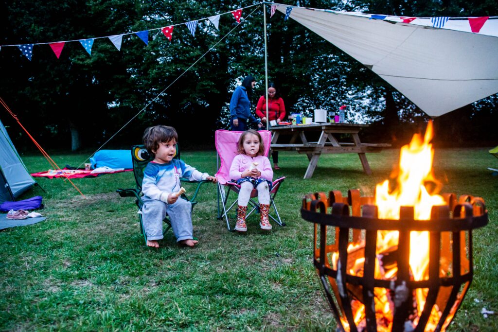 Two young children sitting in camping chairs by a bonfire with parents preparing food in the background at a campsite with bunting decorations