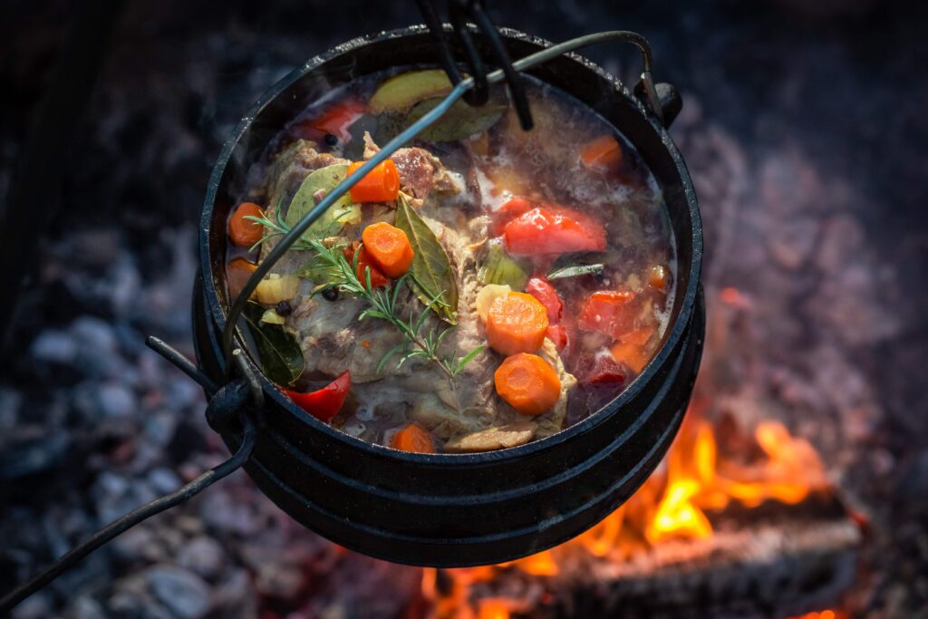 A black cast iron Dutch oven hanging over an open fire, filled with a simmering stew made of meat, carrots, red bell peppers, and fresh herbs, highlighting the outdoor cooking process.