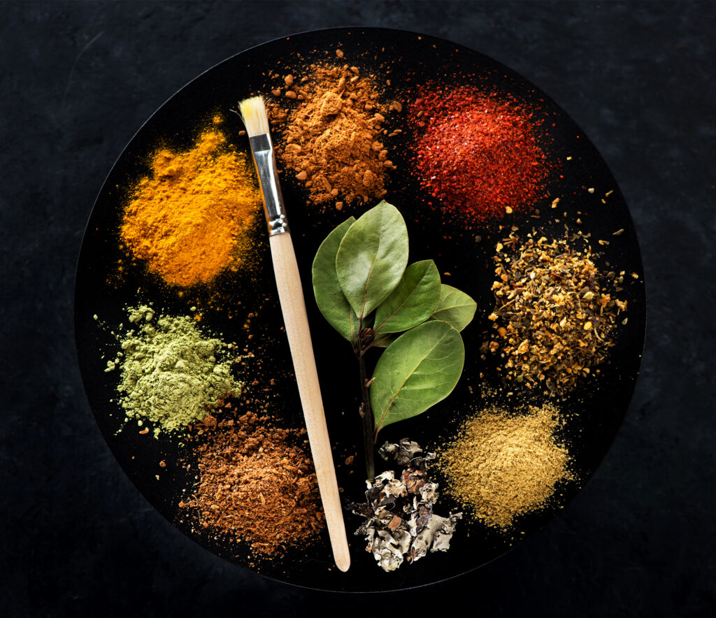 A black plate containing a variety of colorful spices, including turmeric, paprika, and dried herbs, along with fresh bay leaves and a wooden brush, representing the importance of seasoning in cooking.